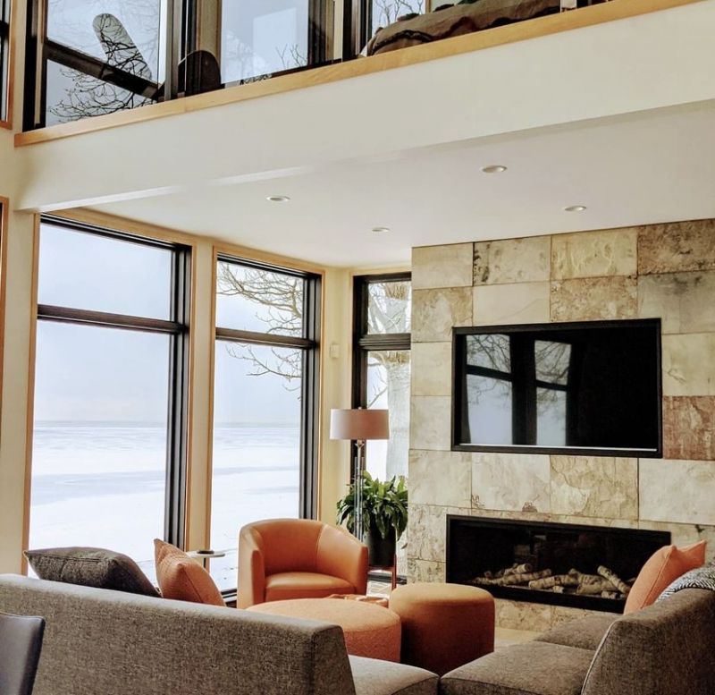 Two-story lakefront great room with floor-to-ceiling windows, a stone fireplace wall and orange accent seating